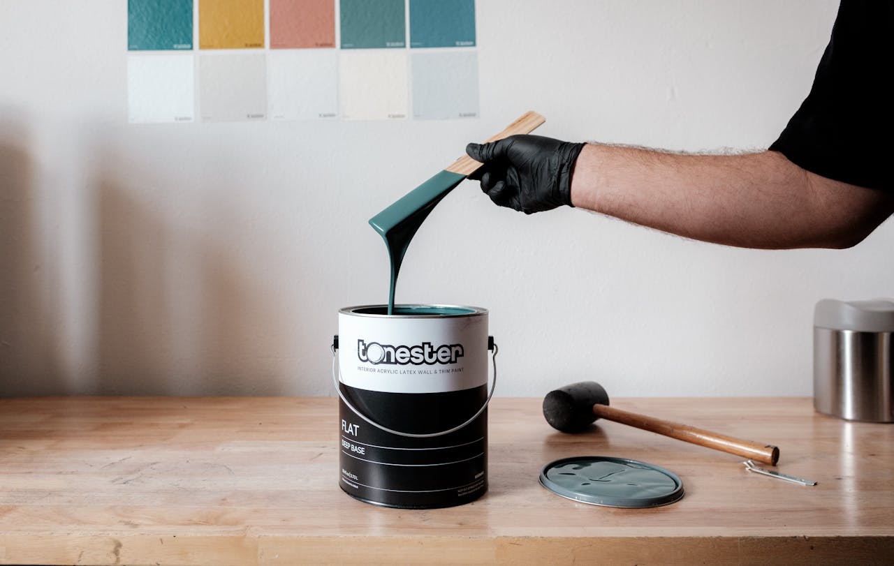 Arm holding a paintbrush over a bucket with green paint, tools on a wooden table.
